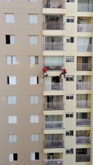 Two painters working in full safety gear, including helmets and harnesses, rappel down the facade of a tall residential building to perform maintenance and painting on the exterior urban wall. 4k.