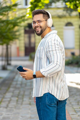 Obraz premium Back view of carefree young man tourist in wireless headphones listening favorite energetic disco music dancing outdoors. Caucasian guy traveler walking spinning on urban city street. Town lifestyle.