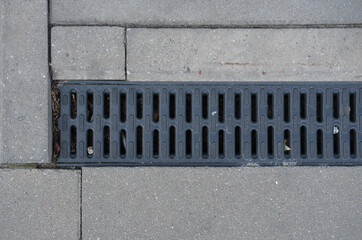 Storm drain grate embedded in the sidewalk, showing a clean surface and slot pattern for water drainage.