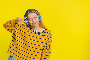 Happy joyful Caucasian woman in blouse listening music on headphones and dancing disco fooling around having fun expressive gesticulating hands. Young adult girl isolated on yellow background indoors