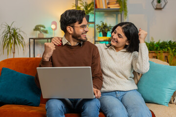 Cheerful exited diverse couple in casual clothes using laptop surprised with email winning lottery online game jackpot on sofa in living room at home. Happy Indian family clenching fists in apartment