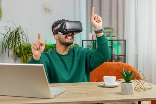 Indian man using virtual reality futuristic technology VR app headset helmet to play simulation 3D 360 online video game, watching film movie at modern home apartment. Guy in goggles sitting at desk