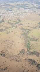 Aerial view with airplane shadow on a sprawling rural landscape featuring rolling hills, dry fields, and scattered vegetation, with a small town visible. High angle perspective of the countryside.