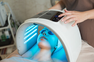 Woman receiving LED light therapy with dome device while beautician adjusts settings