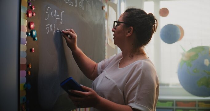Female Teacher Writing on the Chalkboard, Talking to Students, Explaining Method of Multiplying Numbers to Elementary School Children During Math Lesson. Mathematics Class in Modern Colorful Classroom - Powered by Adobe