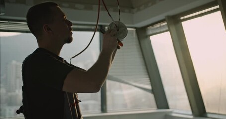 An electrician installs a fire alarm in a modern office building under construction. The sun shines through the window, creating a dramatic silhouette.