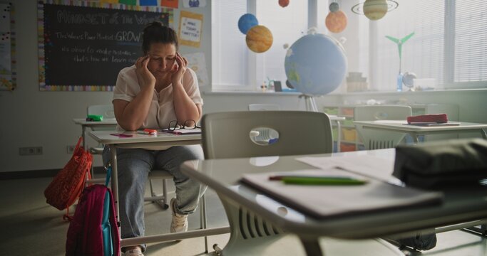 Exhausted Female Teacher Rubbing her Temples, Sitting Alone at Desk in Empty Classroom, Having Rest After Lesson. Woman Feeling Stress, Burnout and Emotional Struggle, Working in Modern Primary School - Powered by Adobe