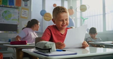 Teacher Giving Papers with School Test Results to Primary School Children Sitting at Desks in Modern Classroom. Smart Young Boy Rejoicing and Celebrating Excellent Grade on Geography or Science Exam.