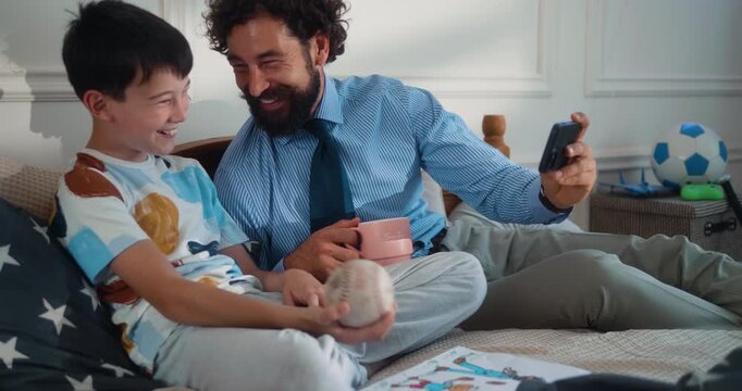 Father in business attire and his son lie on the bed, laughing while watching a phone. Dad enjoys a morning coffee, sharing a warm moment. Part  of the &ldquo;Dad&rsquo;s Day&rdquo; series.