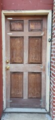 Old wooden front door with peeling varnish, brass handles, and visible wear. Framed by aged trim and red brick wall, showing rustic urban characte