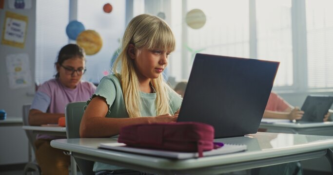 Computer Science Lesson: Primary School Girl Sitting at Desk, Using Laptop in Classroom. Group of Boys and Girls Learning Basic Programming Languages and Internet Safety on Digital Tablets, Laptops.
