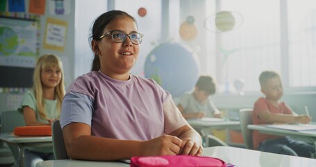 Obraz premium Primary School Girl Sitting at Desk, Raising Hand and Giving Correct Answer on Teacher's Question During Geography Lesson. Group of Young Boys and Girls Learning Science in Modern Colorful Classroom.