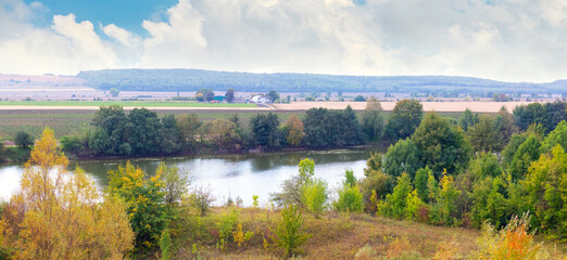 A beautiful landscape with a river and green trees on the bank, single autumn colors on the leaves under a blue sky with clouds