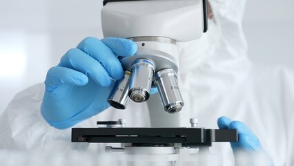 Scientific researcher or virologist wearing protective gear is tuning microscope lenses opposite...