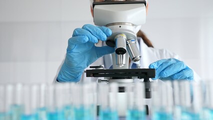 Female scientist wearing protective equipment checking microscope, analyzing blue test tubes in modern laboratory research setting. Medicine, healthcare and science concept