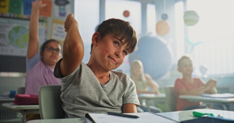 Primary School Boy Sitting at Desk, Raising Hand and Giving Correct Answer on Teacher's Question During Geography Lesson. Group of Young Boys and Girls Learning Science in Modern Colorful Classroom.