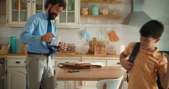 Happy business father and his son start their morning in the kitchen. Dad pours coffee-to-go, son grabs his backpack. Ready for work and school. Part 11 of the &ldquo;Dad&rsquo;s Day&rdquo; series.