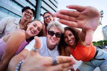 Obraz premium Five cheerful multi-ethnic friends are having fun together outdoors, taking a selfie and making hand frame gestures.