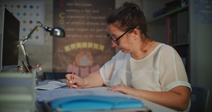 Female Teacher Checking Homework of Students, Grading School Tests After Class, Making Notes. Teacher Sitting at Desk, Working Alone in Empty Classroom, Educating School Subjects in Elementary School.