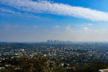 Fototapeta premium A high-angle, panoramic view captures the sprawling Los Angeles cityscape, with the downtown skyline rising in the distance beneath a bright blue, cloud-strewn sky.