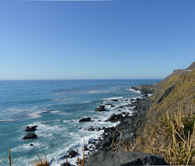 A high-angle view from a grassy cliff overlooking a rugged, rocky coastline where turquoise waves crash ashore under a vast, clear blue sky.