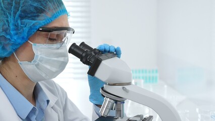 Female scientist analyzing sample through microscope, wearing protective lab gear during medical...