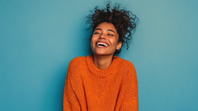 happy emotions concept positive dark skinned beautiful young woman laughs poisitively looks aside with carefree face expression wears casual orange sweater isolated over blue studio background no log