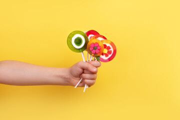 Close-up of a child’s hand holding a bunch of vibrant, fruit-shaped lollipops on sticks against a yellow background.