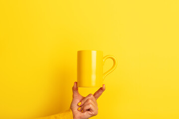 Hand in yellow sweater balancing a plain yellow ceramic mug on fingertips against a bright yellow background.