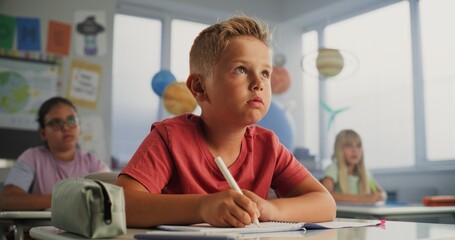 Primary School Boy Sitting at the Desk, Writing Down Lecture in School Notebook. Group of Young Boys and Girls Doing Writing Tasks on Geography or Environmental Science in Modern Colorful Classroom.