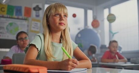Elementary School Girl Sitting at Desk, Doing Writing Tasks in School Notebook. Team of Smart Diverse Kids Writing Down Lecture During Geography Lesson, Studying Science in Modern Colorful Classroom.