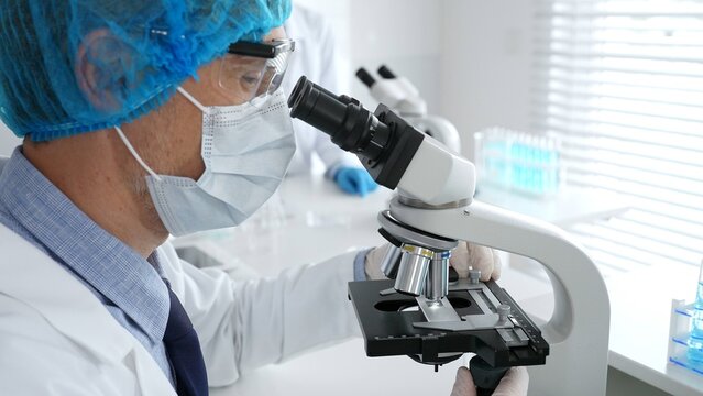 Senior male scientist wearing a protective mask, cap, and gloves, examining samples under a microscope in a modern laboratory. Medicine, healthcare and science concept