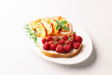 Juicy homemade toasts with cream cheese, raspberry, peach slices and aromatic herbs on the white plate on light stone table close up.