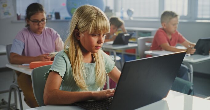 Computer Science Lesson: Primary School Girl Sitting at Desk, Using Laptop in Classroom. Group of Boys and Girls Learning Basic Programming Languages and Internet Safety on Digital Tablets, Laptops. - Powered by Adobe