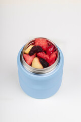 Blue stainless steel insulated food container thermos with fresh mixed fruit pieces watermelon apple dried fruits on white background vertical photo