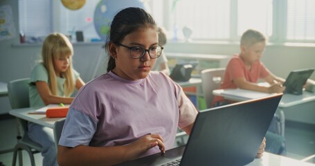 Elementary School Girl Sitting at Desk, Using Laptop in Classroom. Team of Smart Diverse Kids Working on Their Laptops and Tablet Computers, Studying Computer Science, Basic Programming and Coding.