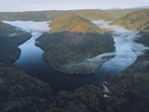 Aerial view of the Sil River winding through the lush, forested hills, with ribbons of fog clinging to the valleys, O Savinao, Lugo, Spain.