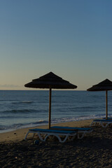Two empty sun loungers and a straw umbrella sit on a sandy beach, with a calm ocean and distant mountains under a clear sky.