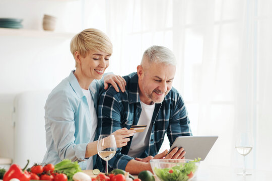 Healthy eating in middle age, paying bills, ordering groceries and shopping online. Smiling senior couple use laptop and credit card in modern comfort kitchen interior with bright colored vegetables
