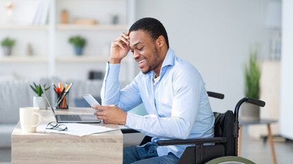 Remote job for with disability people. Happy black guy in wheelchair communicating online, using smartphone and laptop indoors. African American man in taking online course, browsing web