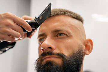 A barber is using clippers to create a trendy haircut on a man with a full beard, who looks relaxed in a contemporary barbershop atmosphere