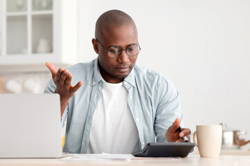 Savings and finances concept. Shocked black mature man calculating utility charges and bills, using calculator and laptop computer, sitting in the kitchen at home. Man having financial problem