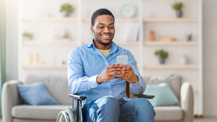 Black man in wheelchair using smartphone, browsing web or watching movie at home. Young guy with disability checking social media, speaking to friend on mobile device