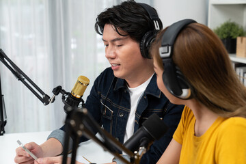 Smiling radio influencer hosts wearing headphones with channel social media online live streaming with talking topic on script note and laptop to listeners at morning time at studio record. Infobahn.