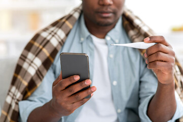 Consultation with doctor. Unrecognizable sick black man texting on phone measuring body temperature, having fever, wrapping in blanket. Covid-19, cold and influenza symptom.