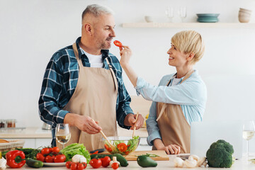 Useful eating in middle aged, vegetarianism and health care at home. Happy adult man and woman have fun, lady feeding man piece, preparing salad in kitchen interior with fresh vegetables on table