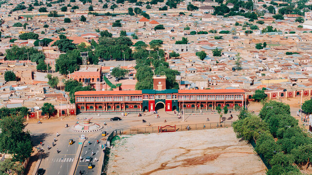Aerial view of the expansive, terracotta-roofed building with its stately clock tower, framed by lush green trees and the dusty landscape, Katsina, Katsina, Nigeria.