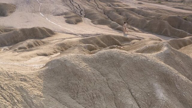 Mysterious man wearing a white robe and holding a wooden staff in his hand, walking through the white, sandy hills of a mountain. Aerial dolly out shot