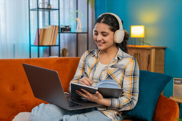 Indian young woman listening to music on laptop with headphones, carefully writing notes in notebook. Arabian girl focused on freelance work at home, thoughtfully working in comfortable atmosphere.