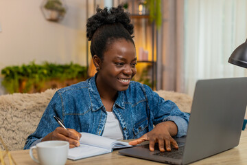 African American young woman studies online at laptop while sitting at table on home sofa. Black girl takes notes in notebook looking focused, attentive, and fully engaged in distance learning process